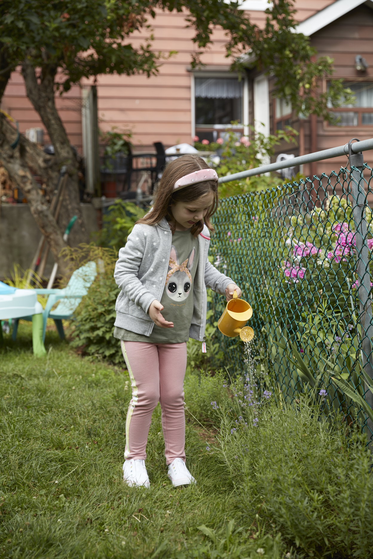 Young girl wearing pink leggings and a hoodie watering plants in backyard 