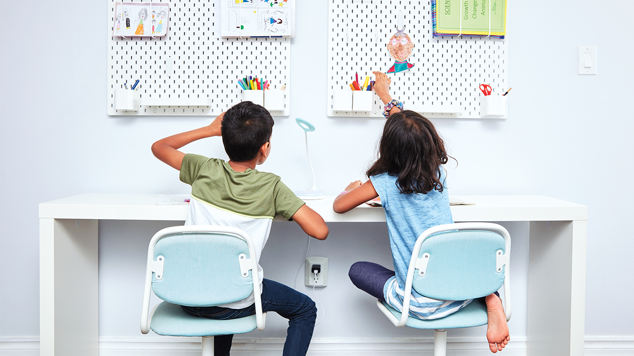 photo of two kids sitting at a desk reaching for pencils that are hung up on the wall