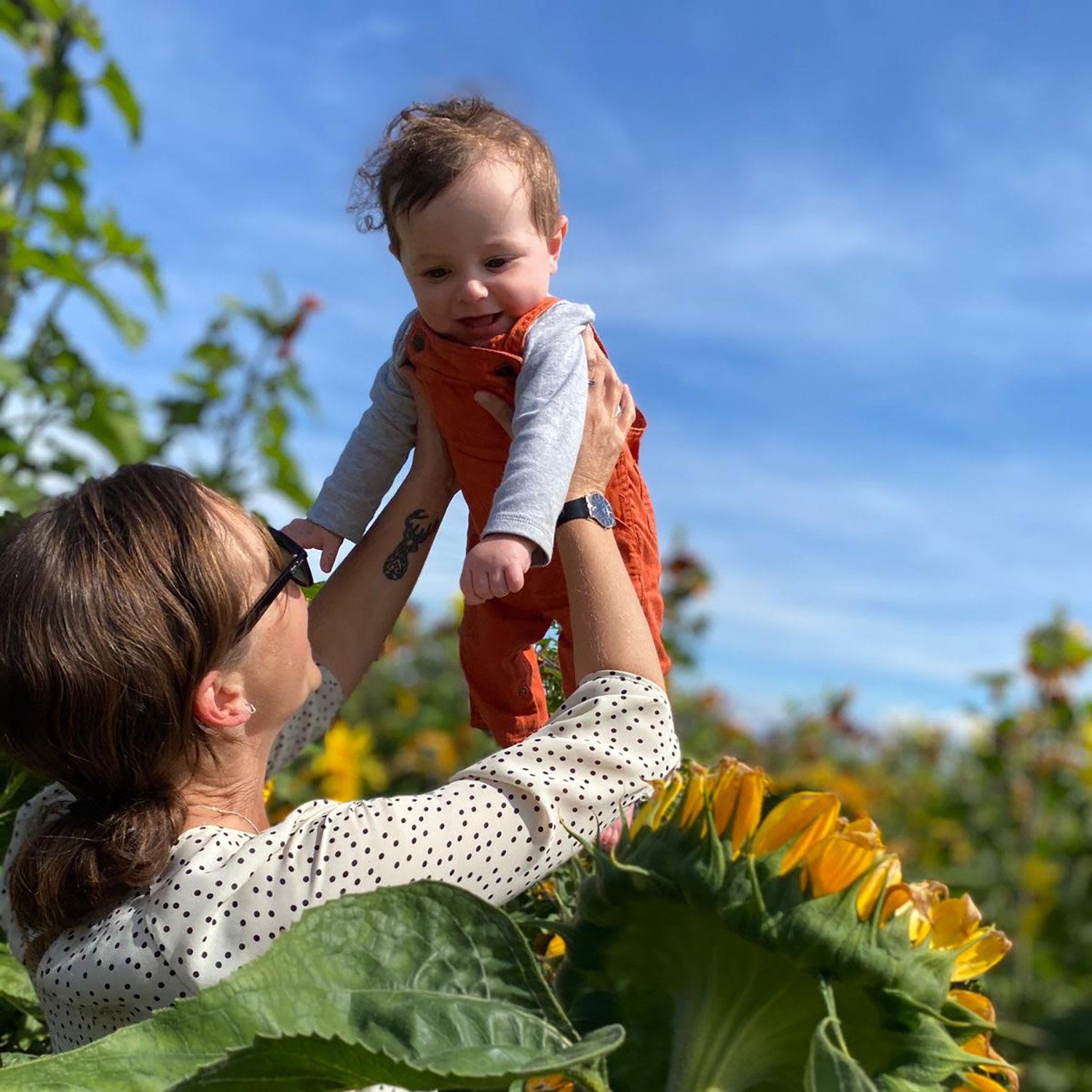 a mom holds her baby up to the sky in a patch of sunflowers