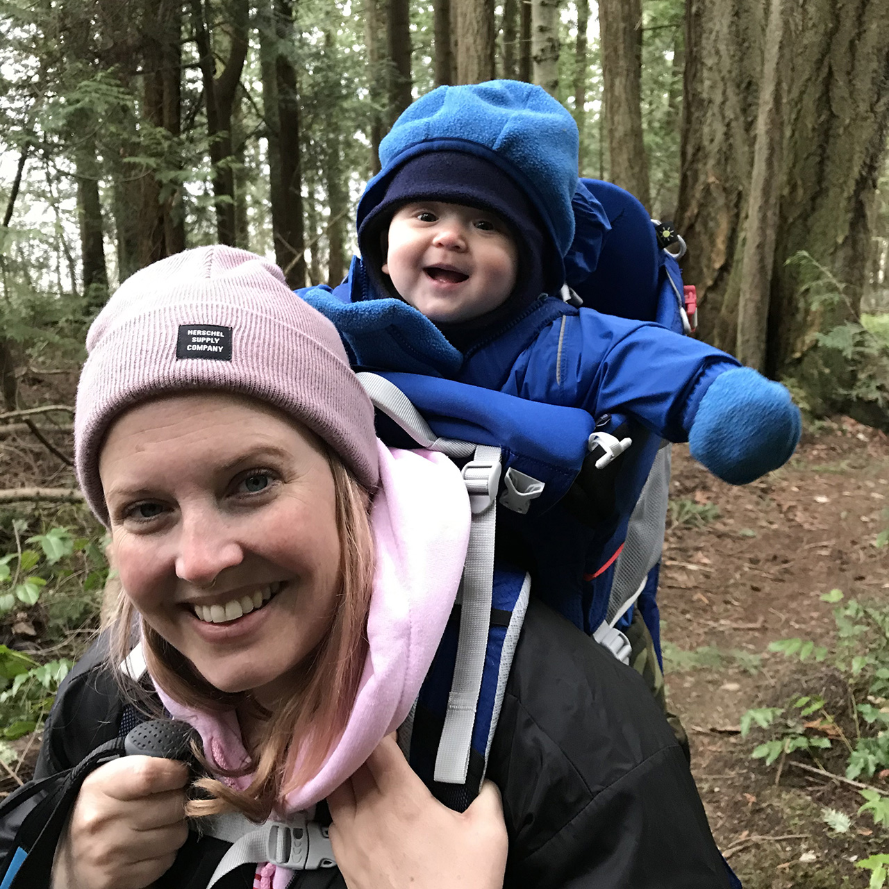 a mom in the woods with her smiling baby in a backpack on her back